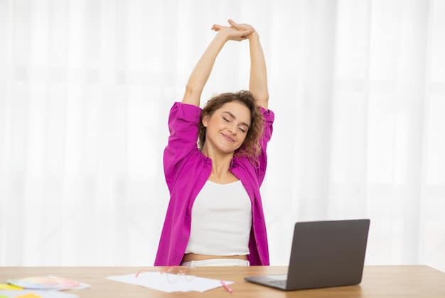 Office worker doing stretching exercises next to her desk and computer. Her hands are above her head, reaching for the sky, and she is smiling with eyes closed.