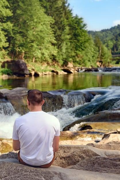 A person meditating outdoors in a serene environment with a waterfall in the background. Their posture is relaxed and their eyes are gently closed.