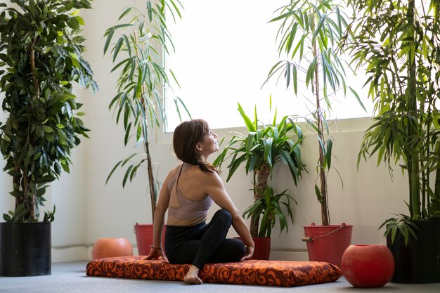 A person practicing yoga on a mat in a serene, sunlit room. The focus is on their peaceful expression and relaxed posture. Plants are visible in the background.