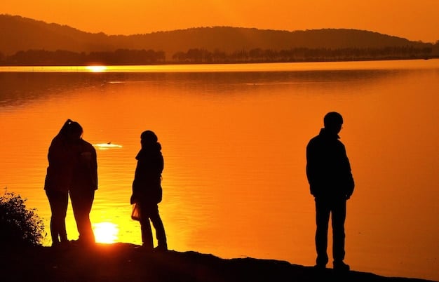 A person silhouetted against a sunset, gazing out at mountains and a tranquil lake. They are holding a journal and pen, seemingly disconnected from electronic devices and fully immersed in the natural environment.