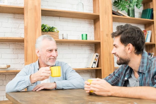 An elderly man with white hair sitting and talking to his son. Both men are smiling and sitting at a table.